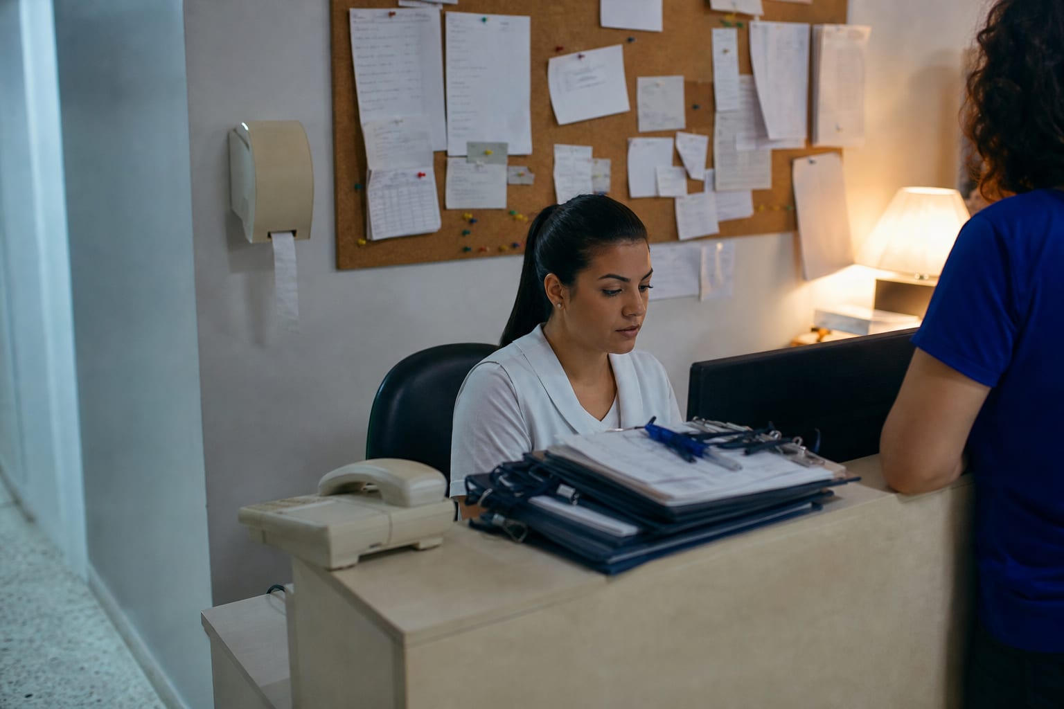 Recepcionista de clínica digita no computador atrás do balcão, com telefone e agenda de papel ao lado